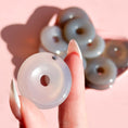 Load image into Gallery viewer, Fingers holding a pale grey agate donut with a pile of donuts in the background on a soft pink background