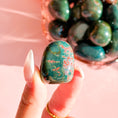 Load image into Gallery viewer, Fingers with white nails hold blue and green bloodstone tumble in the center of the image with an orange bowl full of bloodstone tumbles in the right corner of the image in the background.