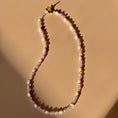 Load image into Gallery viewer, A white and purple beaded necklace lays in the center of the image on a tan background.
