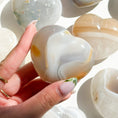 Cargar la imagen en la vista de la galería, Fingers hold a soft grey-brown agate heart with a quartz inclusion with six agate hearts scattered across a white background.