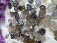 Load image into Gallery viewer, Pile of grey and brown agate mushrooms lay on a white background with purple flowers in the left corner.