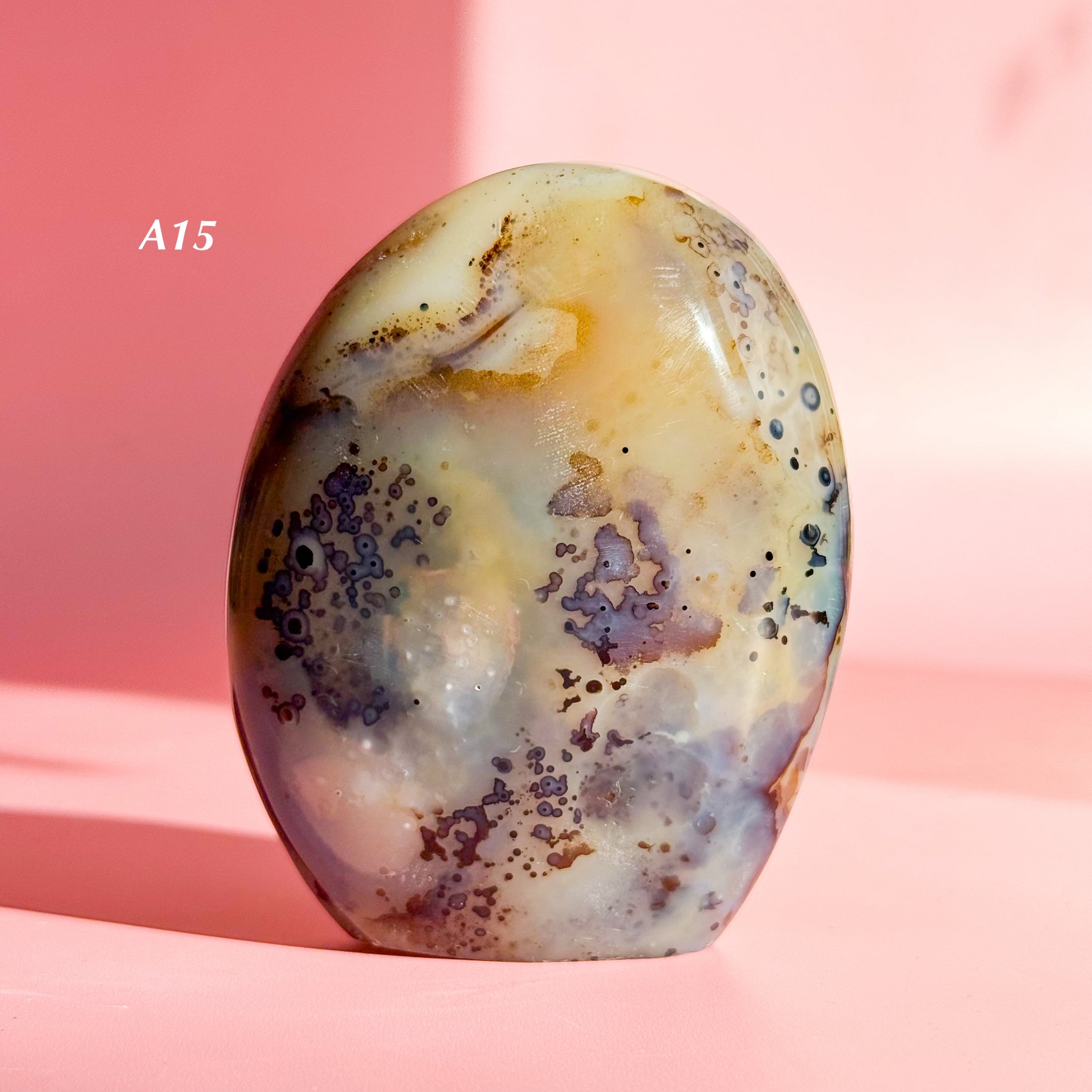 Grey and brown agate freeform with orange inclusions and large black speckles throughout set on a pale pink background. 