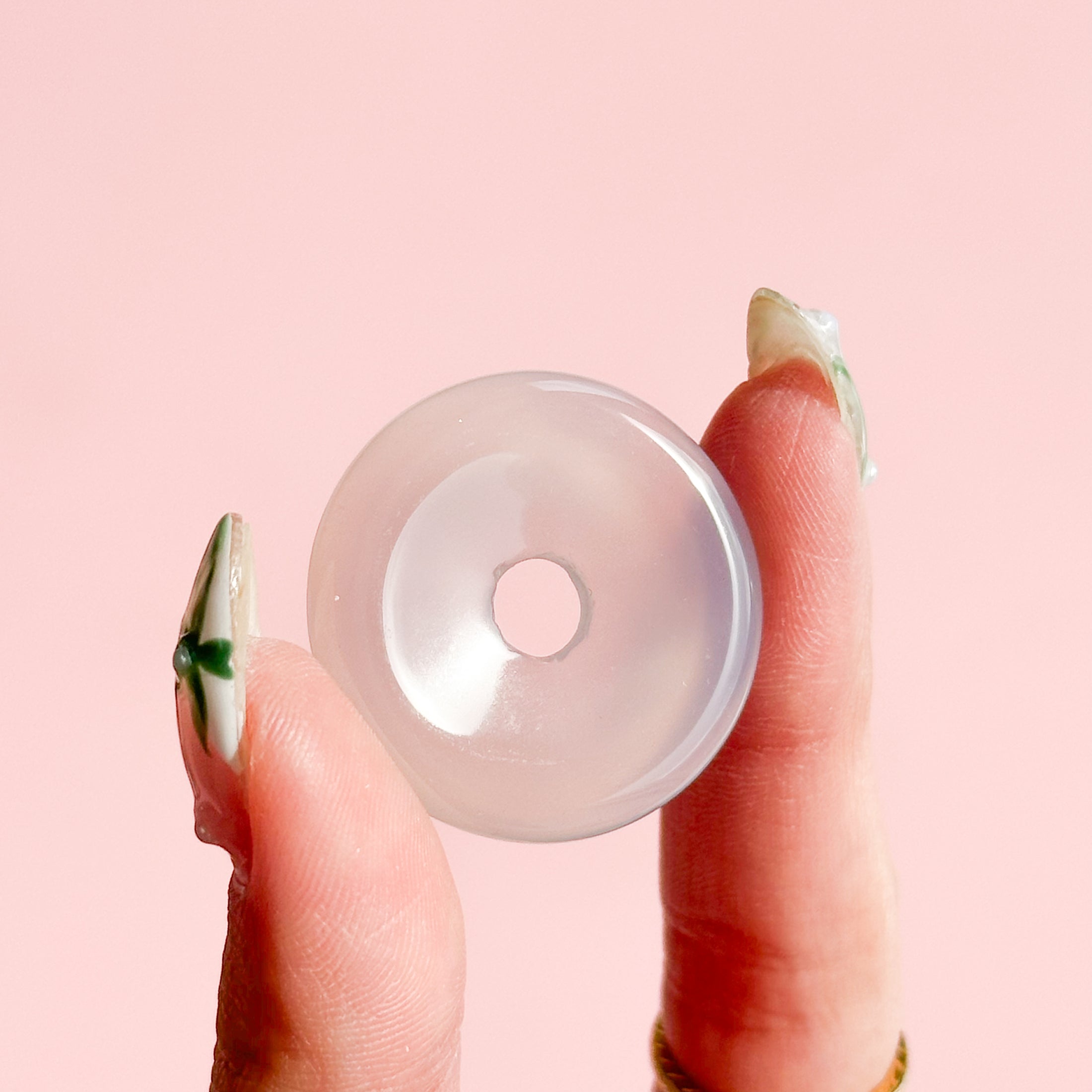 Fingers holding a pale white agate donut on a pale pink background. 