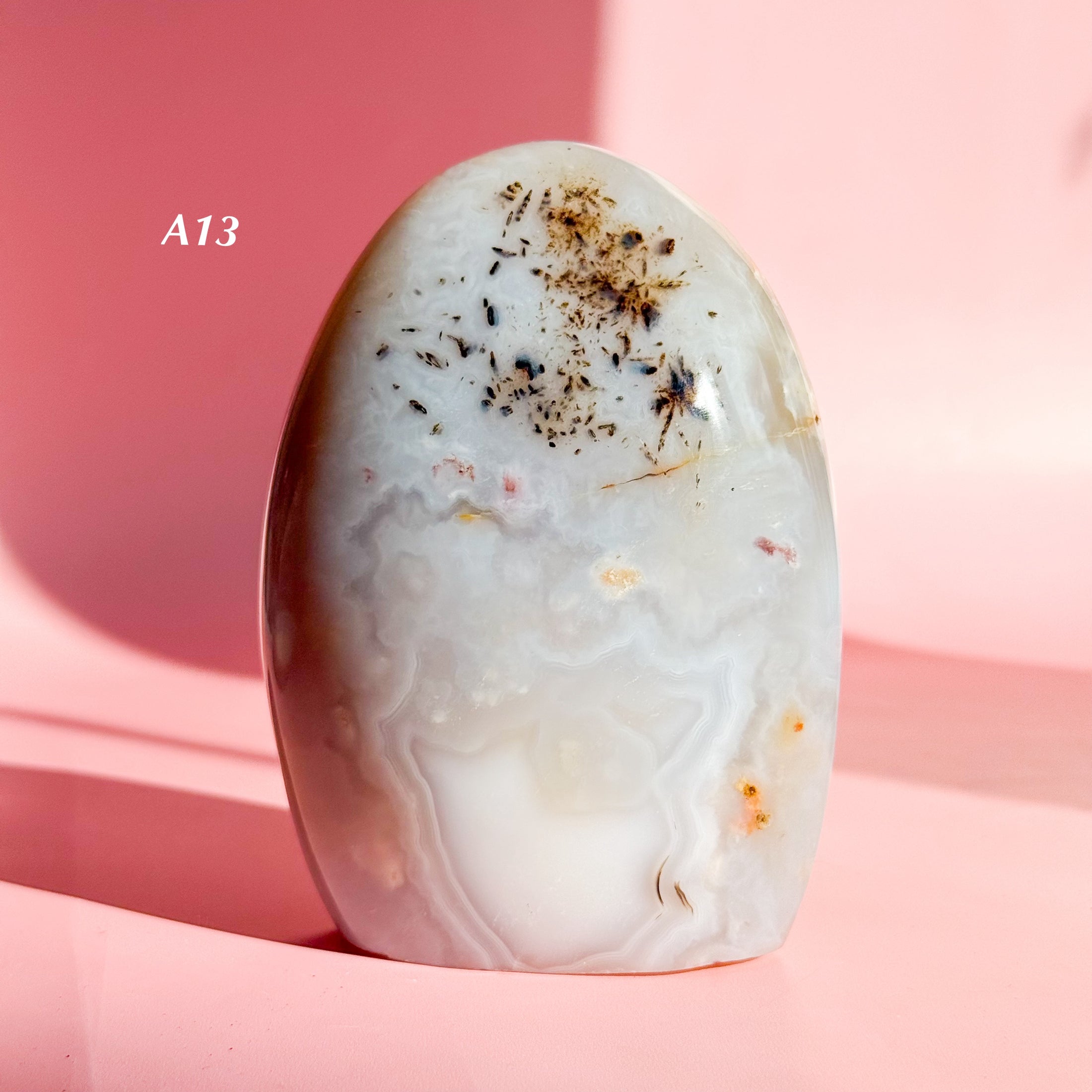 White agate freeform with soft grey inclusions and brown-black speckles on a pale pink background. 