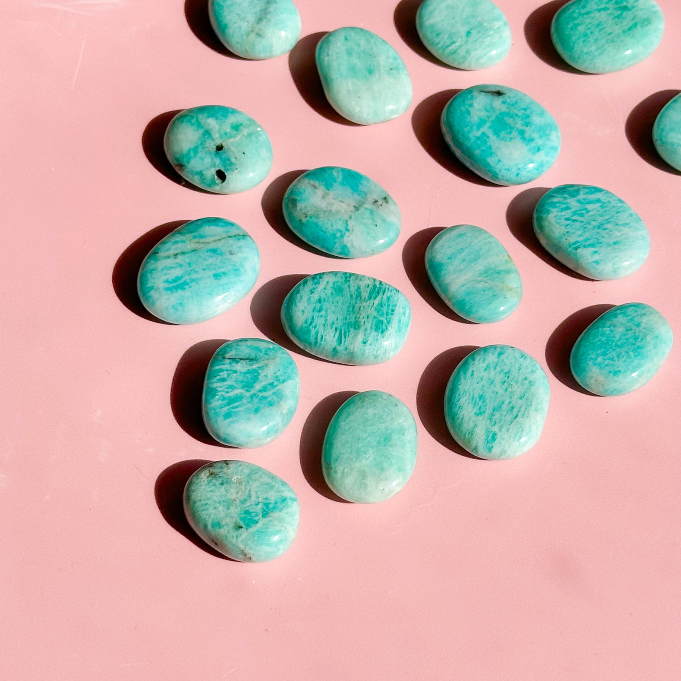 seventeen teal amazonite flat stones lay flat on a pale pink background starting from the top right corner to create a point near the bottom left corner of the image. 