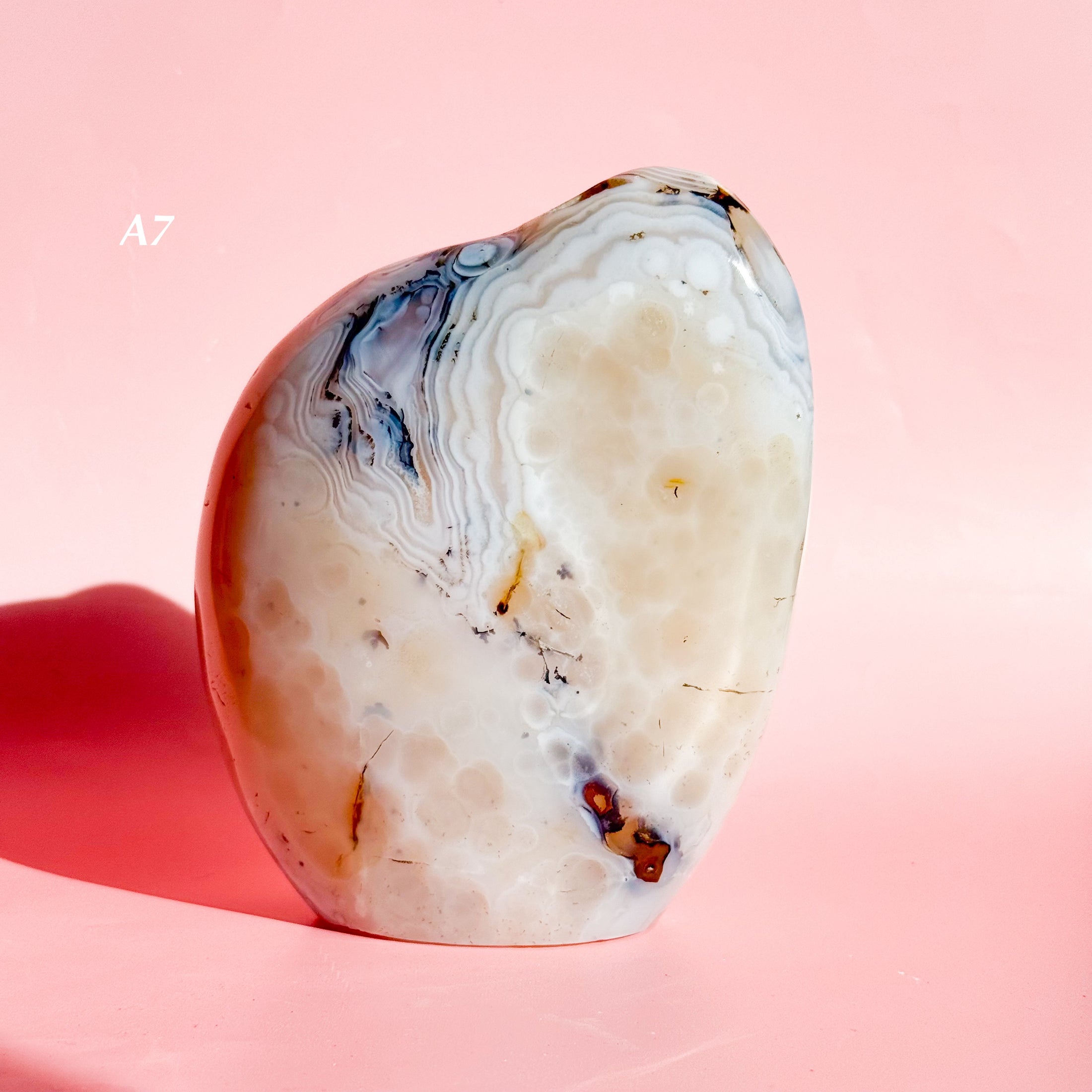 Soft white agate freeform with brown flower inclusions and black speckles throughout, set on a pale pink background. 