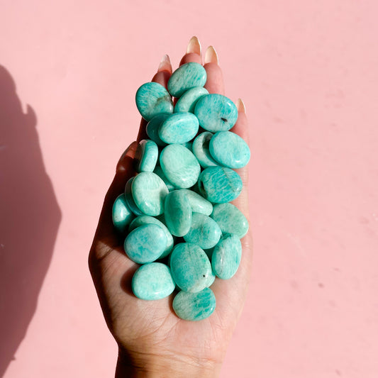 Hand holds a pile of teal amazonite flat stones on a soft pink background. 