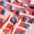 Load image into Gallery viewer, Fingers hold blue angelite tumble in the center of the image with the background full of scattered tumbles 
