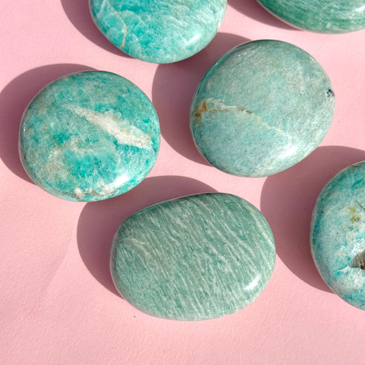 Six teal amazonite palm stones lay flat against a soft pink background. 