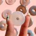 Load image into Gallery viewer, Fingers holding a soft white agate donut with a group of donuts in the background on a soft pink background.
