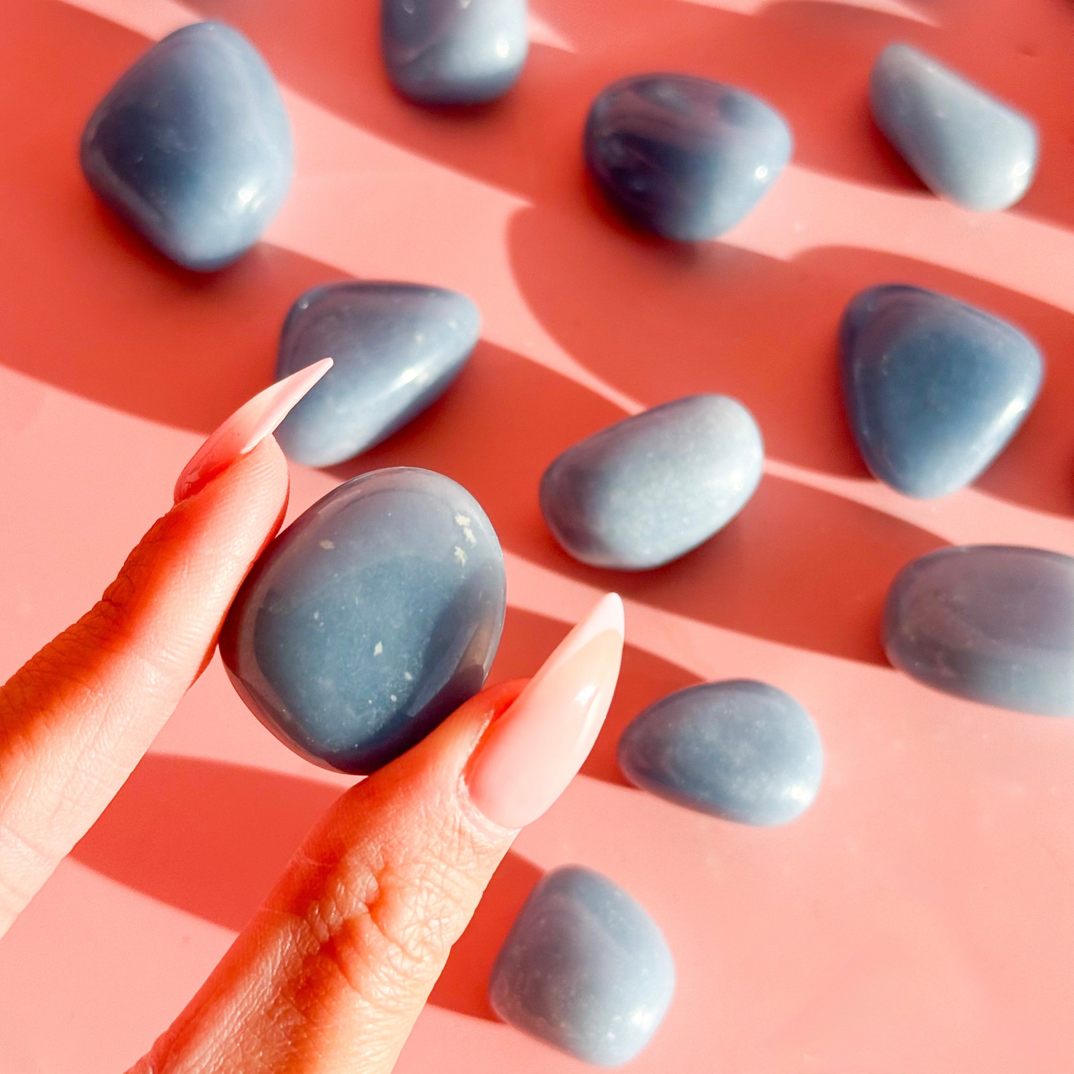 Fingers hold blue angelite tumble with eleven stones lying in the background of the image out of focus