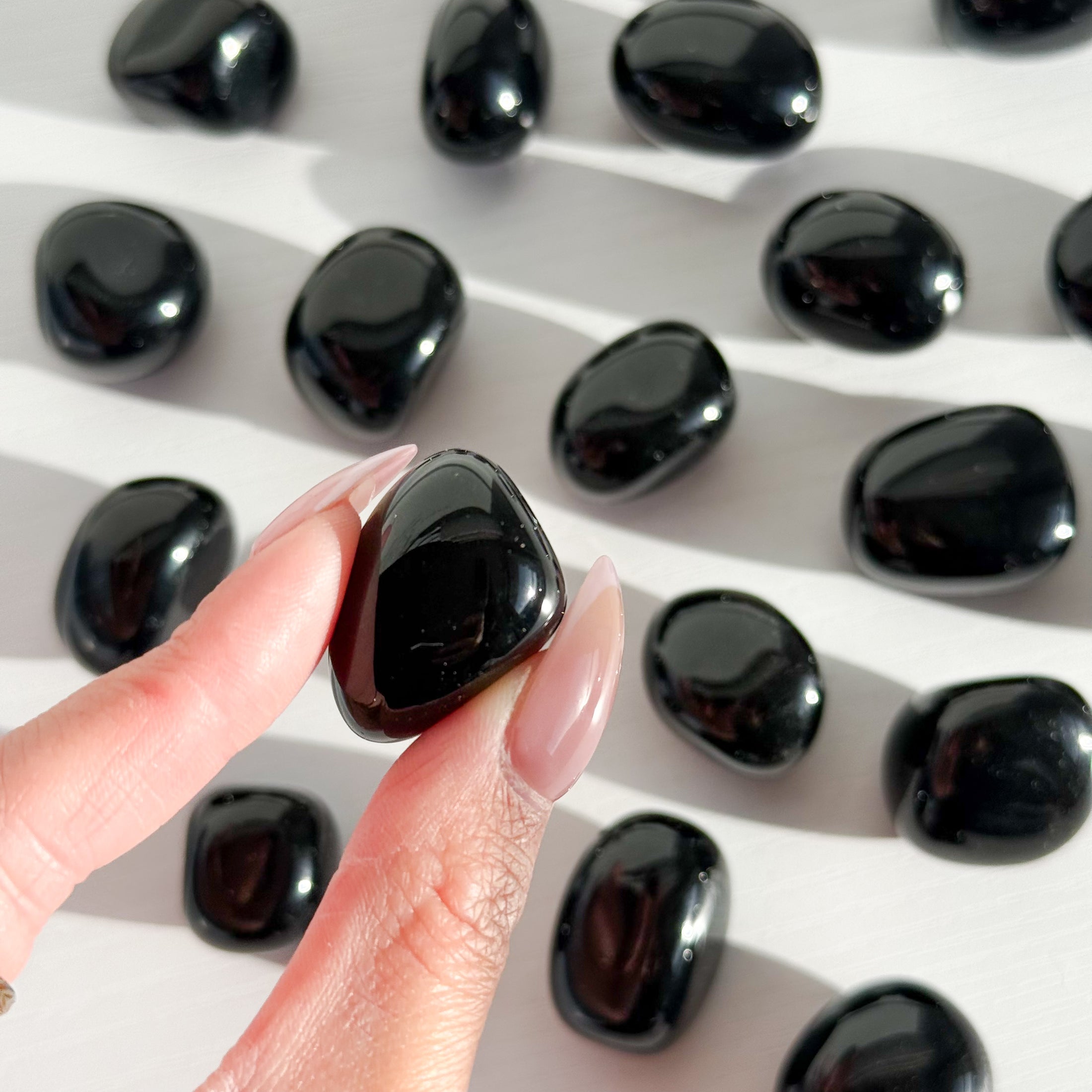 Fingers with pink nails hold one small black obsidian tumble with multiple scattered across the background out of focus. 