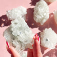 Load image into Gallery viewer, Fingers with red nails hold a white apophyllite cluster in the center of the image, with four clusters out of focus on the right-hand side of the screen on a pink background. 
