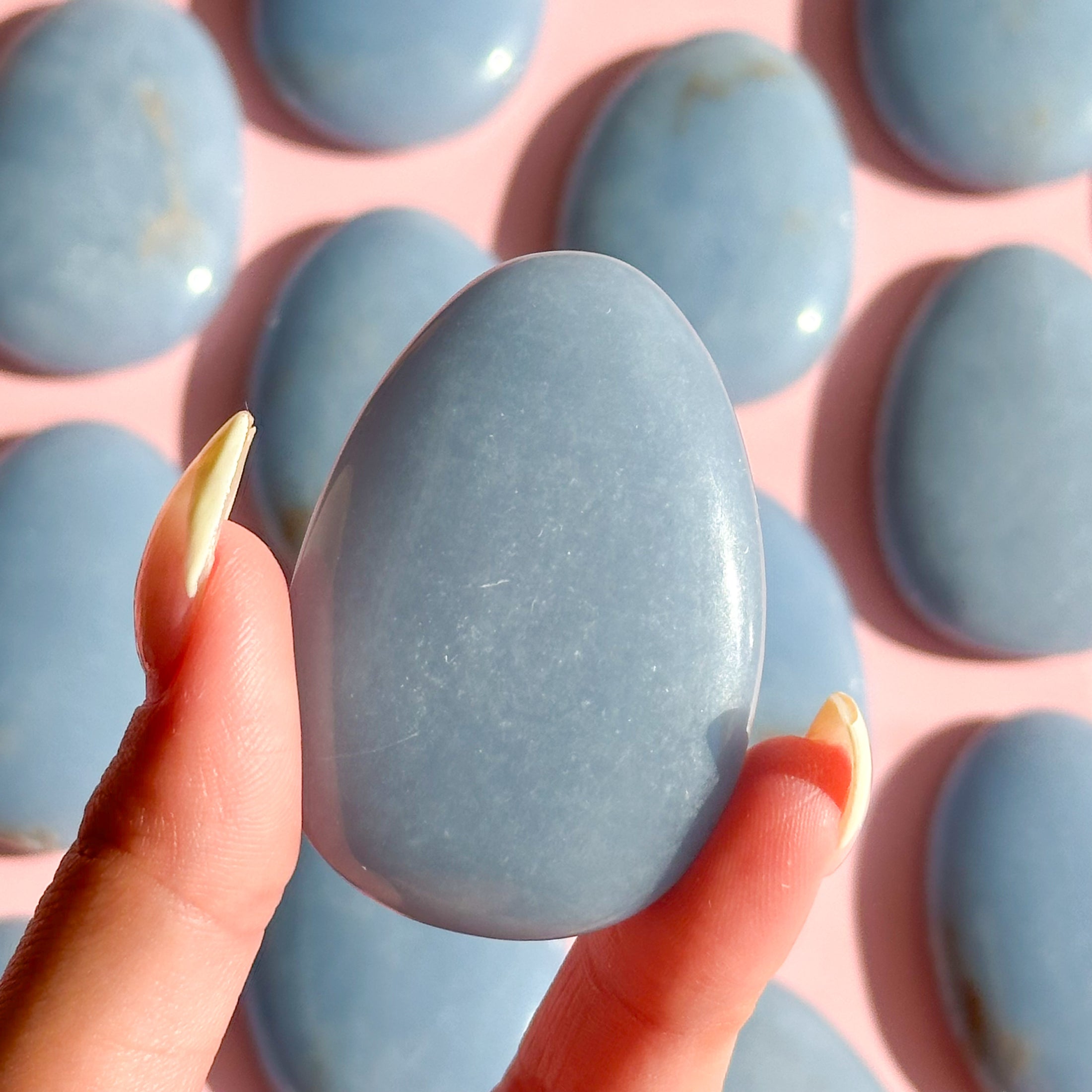 Fingers with yellow nails hold a small, blue angelite worry stone in the center of the image with a group of stones across the background out of focus