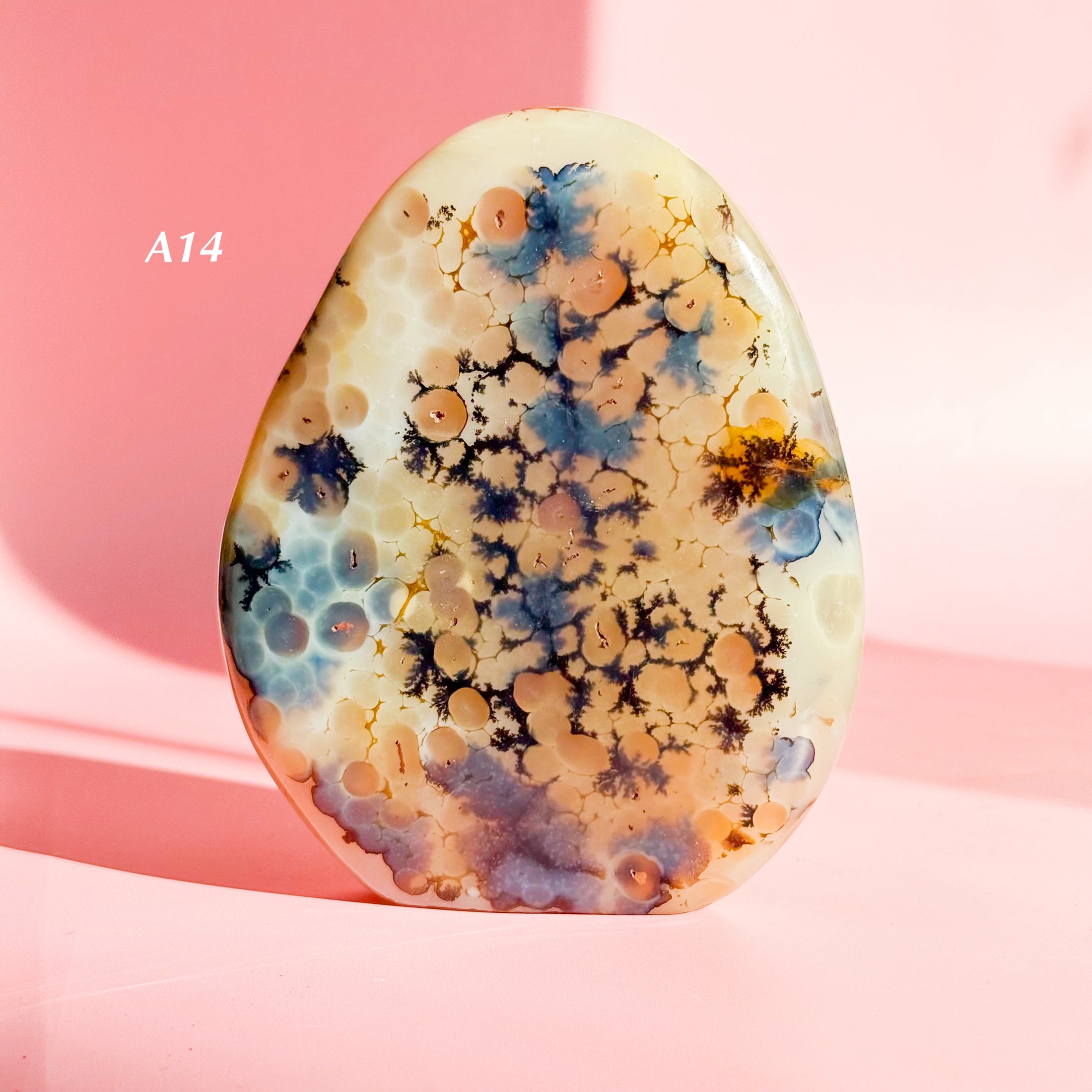 Orange-brown agate freeform with large flower inclusions and black bubbles set on a pale pink background. 