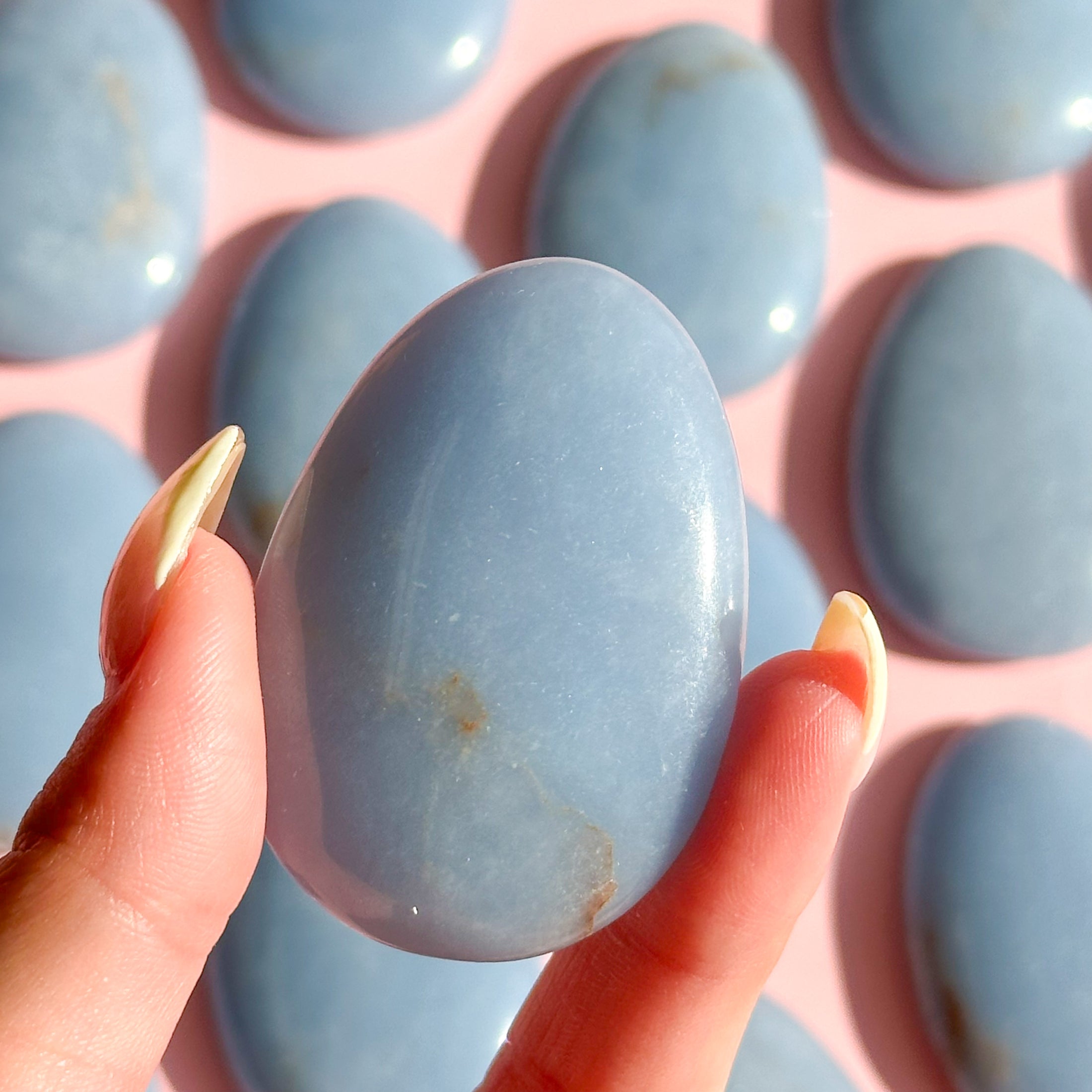 Fingers with yellow nails holds light blue angelite worry stone in the center of the image with eleven stones out of focus in the background of the image. 