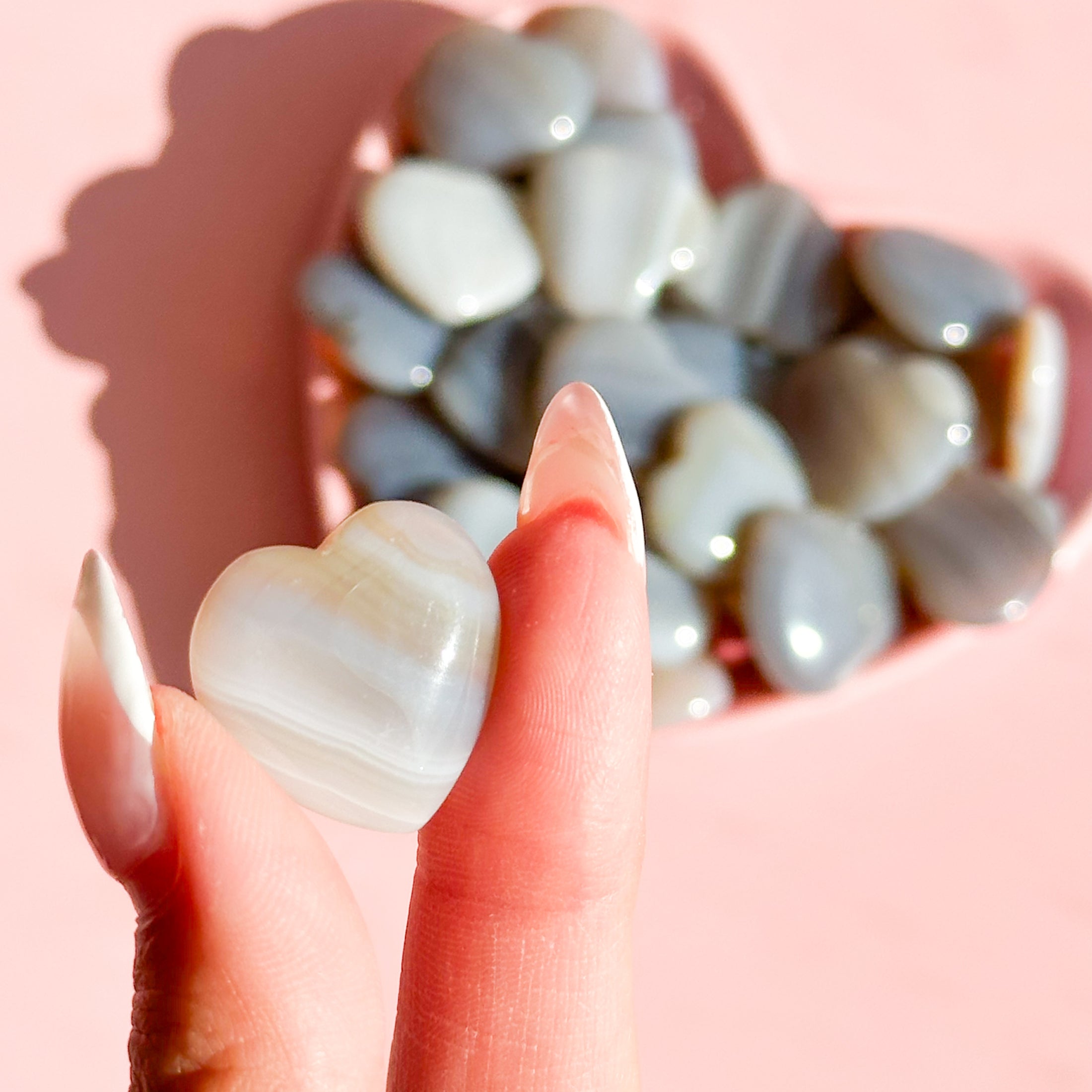 Fingers hold tiny white-brown agate heart with a bowl of agate hearts in the background. 