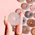 Load image into Gallery viewer, Fingers holding a pale white agate donut with a group of donuts arranged on the far right end of the image on a soft pink background. 
