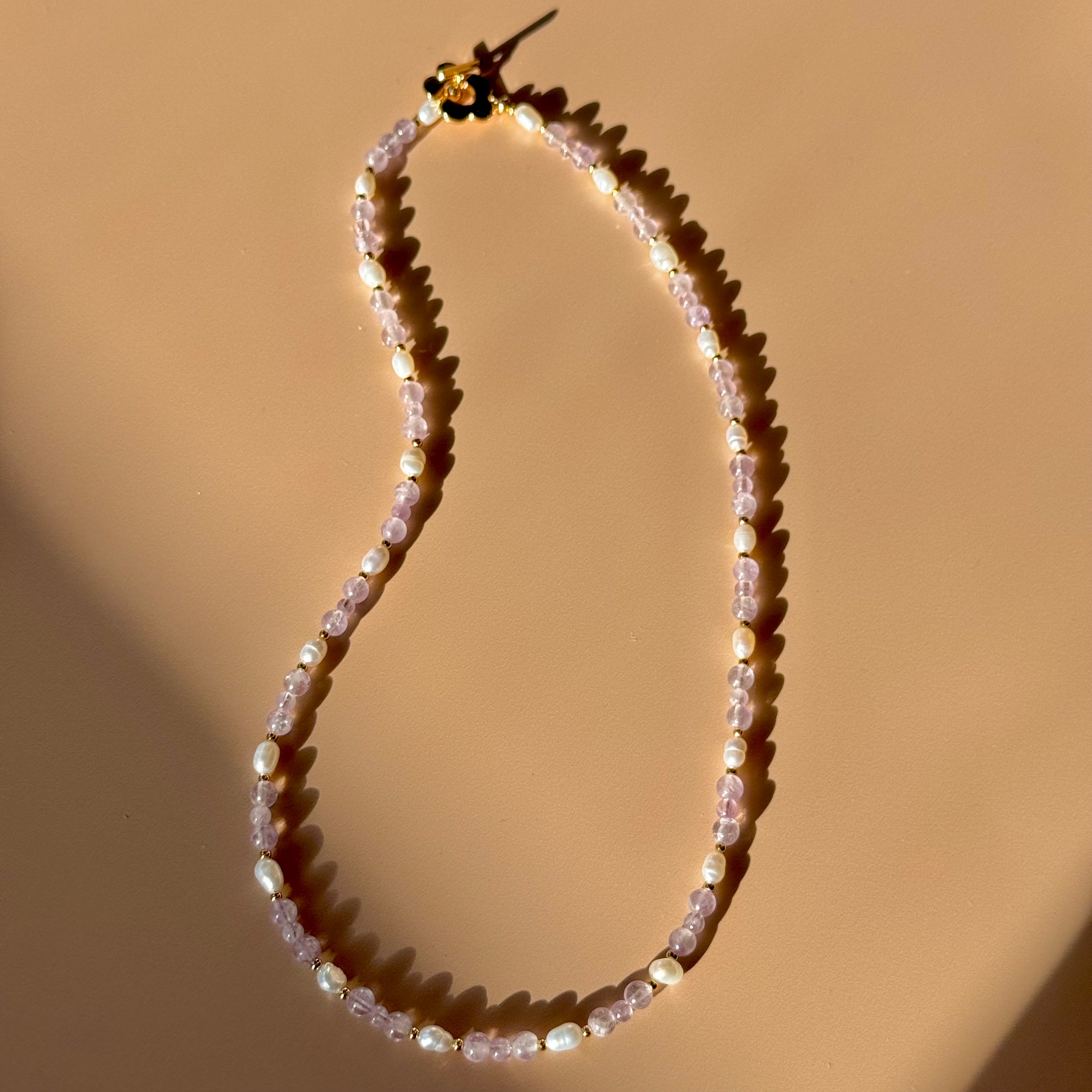 A white and purple beaded necklace lays in the center of the image on a tan background. 