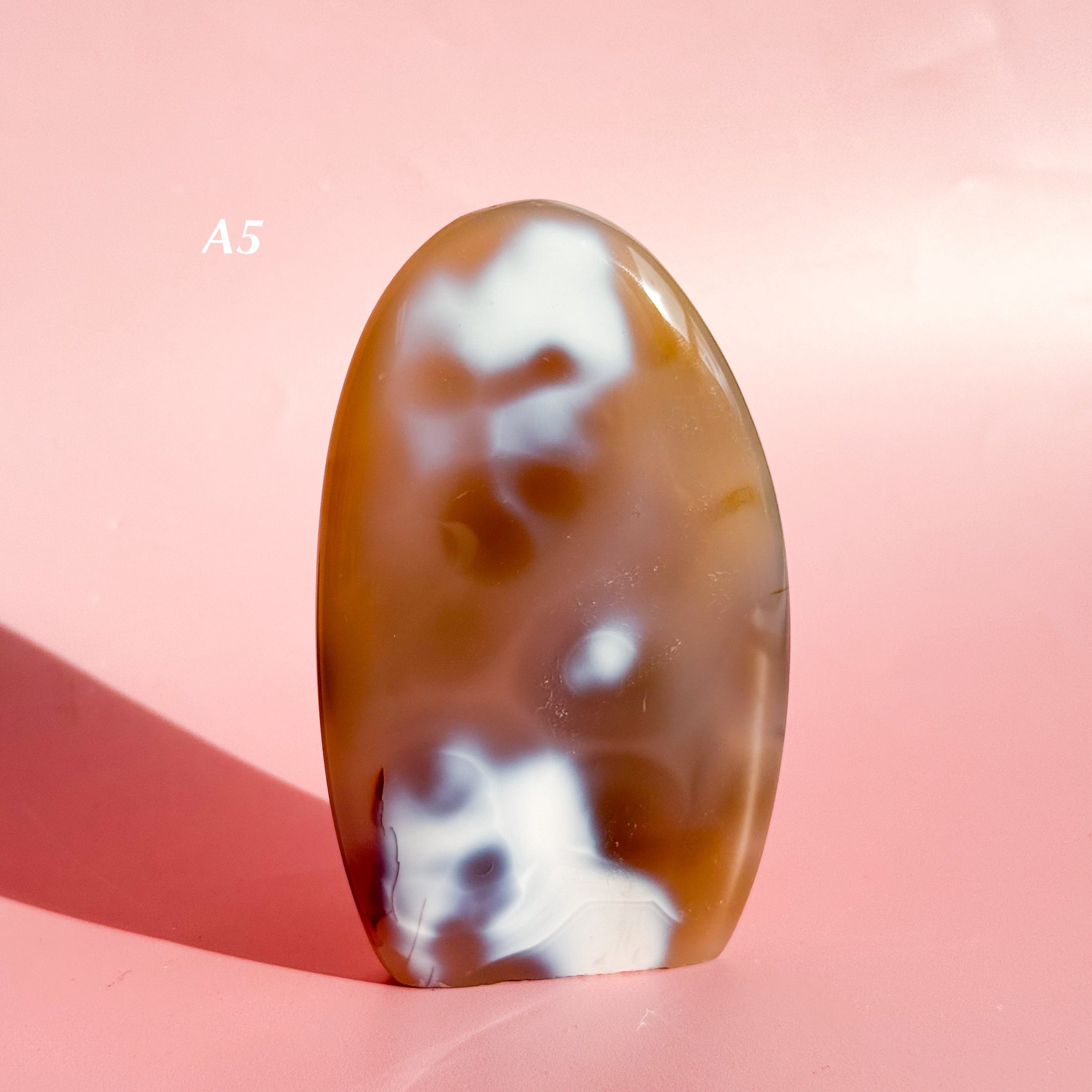 Dark brown-orange agate freeform with clusters of white inclusions on a pale pink background. 