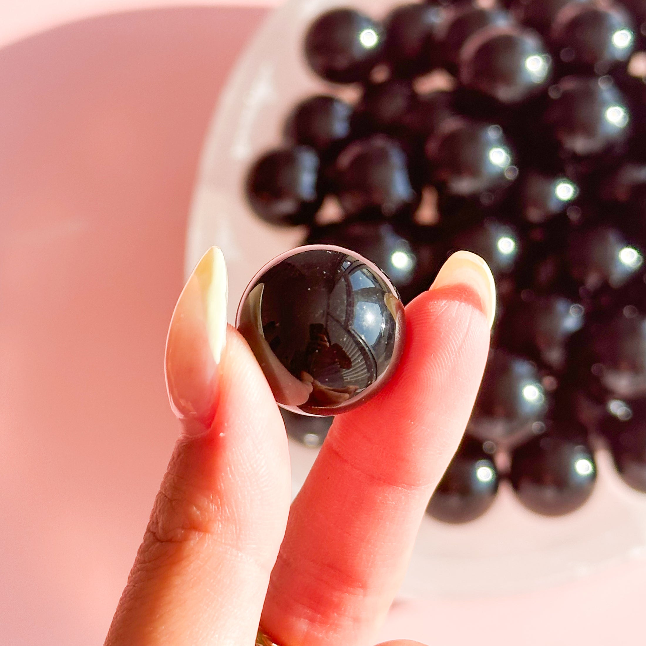 Fingers with yellow nails hold small black obsidian mini sphere with a large bowl of spheres in the background. 