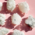 Load image into Gallery viewer, Eight white and grey apophylite clusters lay scattered on the screen on a pink background. 
