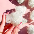 Load image into Gallery viewer, Fingers with red nails hold a white apophyllite cluster in the center of the image, with four clusters out of focus on the right-hand side of the screen on a pink background. 
