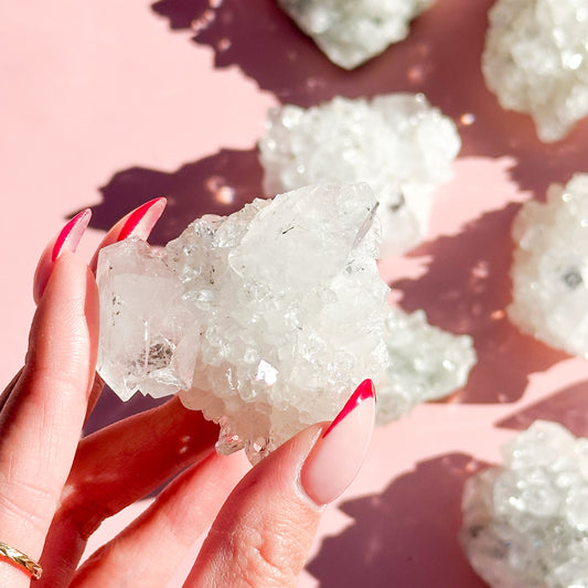 Fingers with red nails hold a white apophyllite cluster in the center of the image, with four clusters out of focus on the right-hand side of the screen on a pink background. 