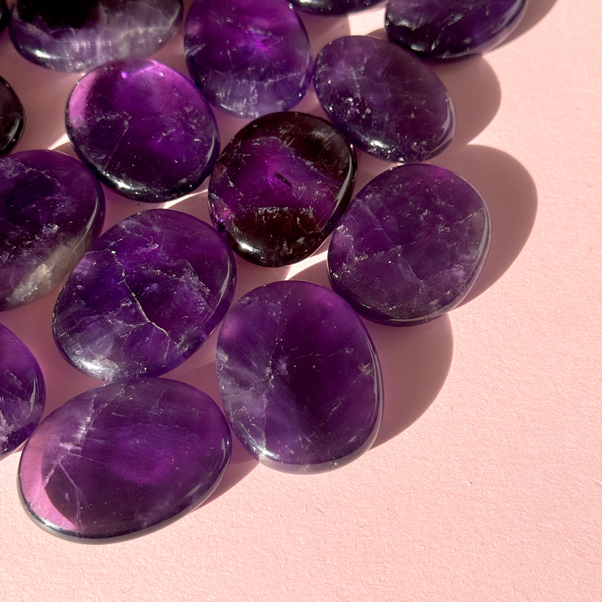 Cluster of dark purple amethyst flat stones lay in the top left corner of the image on a pink background. 