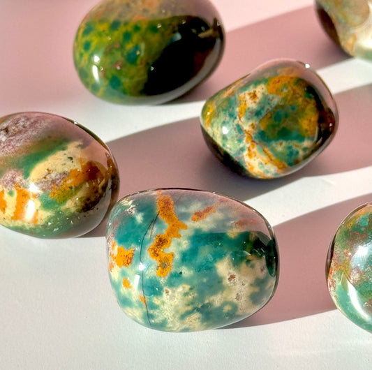 Six green, yellow, and blue bloodstone tumbles lie scattered in the center of the screen on a soft pink background. 
