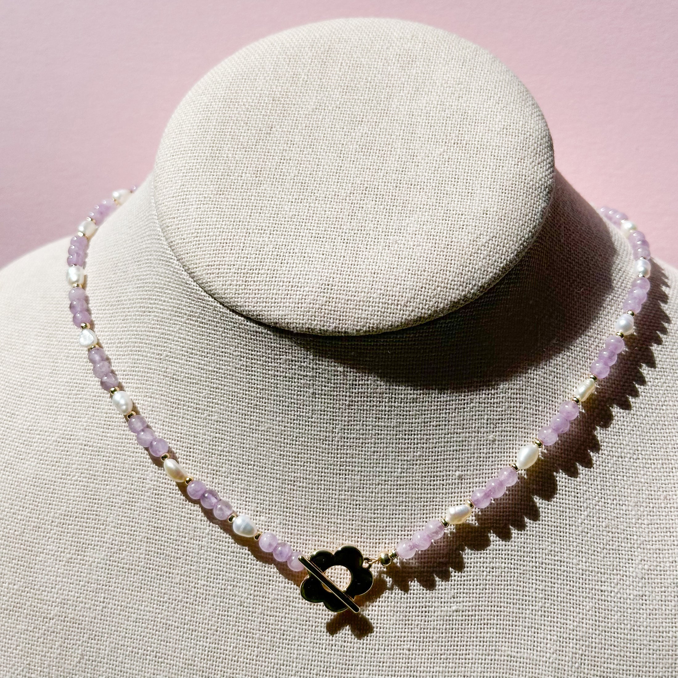 A purple and white beaded necklaced with a gold flower claps sits on a mannequin in the center of the image. 
