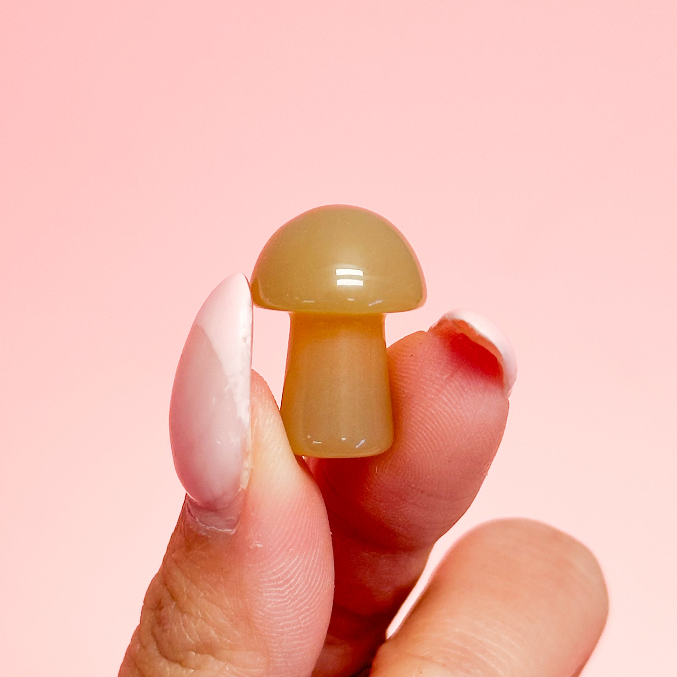Fingers with long white nails holding an orange-brown agate mushroom on a soft pink background. 